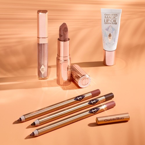 Lip gloss in a nude golden-brown shade in a glass tube with a gold-coloured lid, open nude peachy-brown lipstick in a gold-coloured tube, lip oil in a white-coloured tube with a reflective, geometrical pattern on the front, and three lip liners in nude pink, brown, and nude red shades.