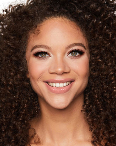 A medium-tone model with green eyes wearing smokey brown and gold eye makeup with warm pink blush and glossy nude-pink lips