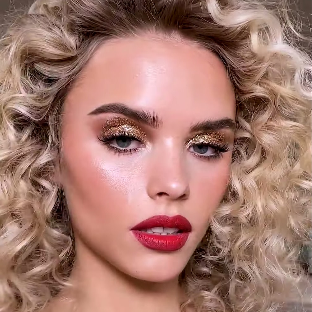 Fair-tone blonde model with blue eyes wearing shimmery gold eye makeup with soft peach blush, highlighter on the high points of her face, and bold, red lipstick. 