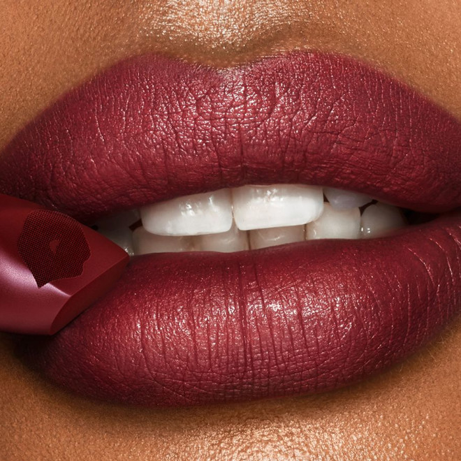 Lips close-up of a deep-tone model wearing a winter berry red, matte lipstick while holding the lipstick in front of her lower lip.