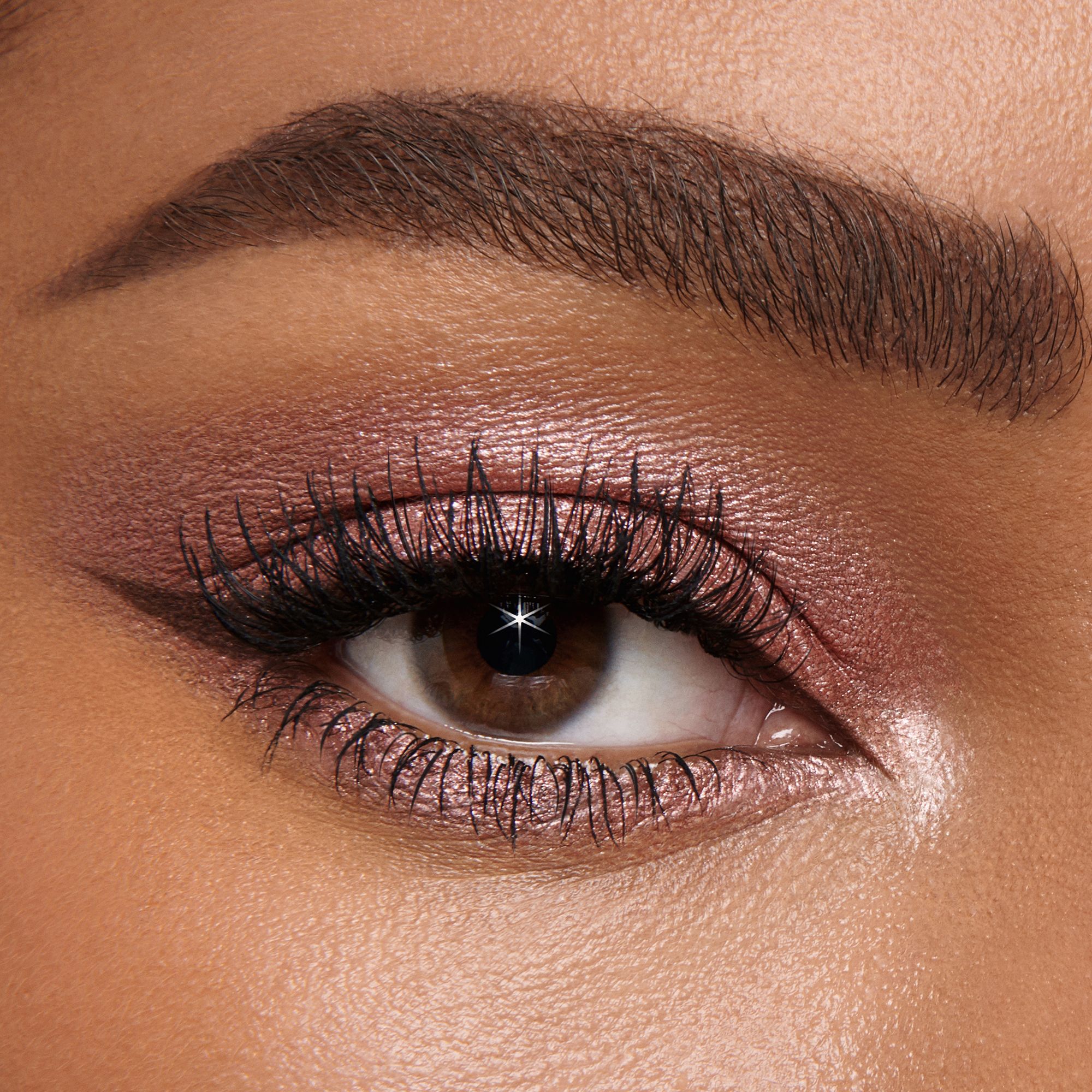 Single-eye close-up of a deep-tone model with brown eyes wearing shimmery rose gold eyeshadow with black eyeliner. 