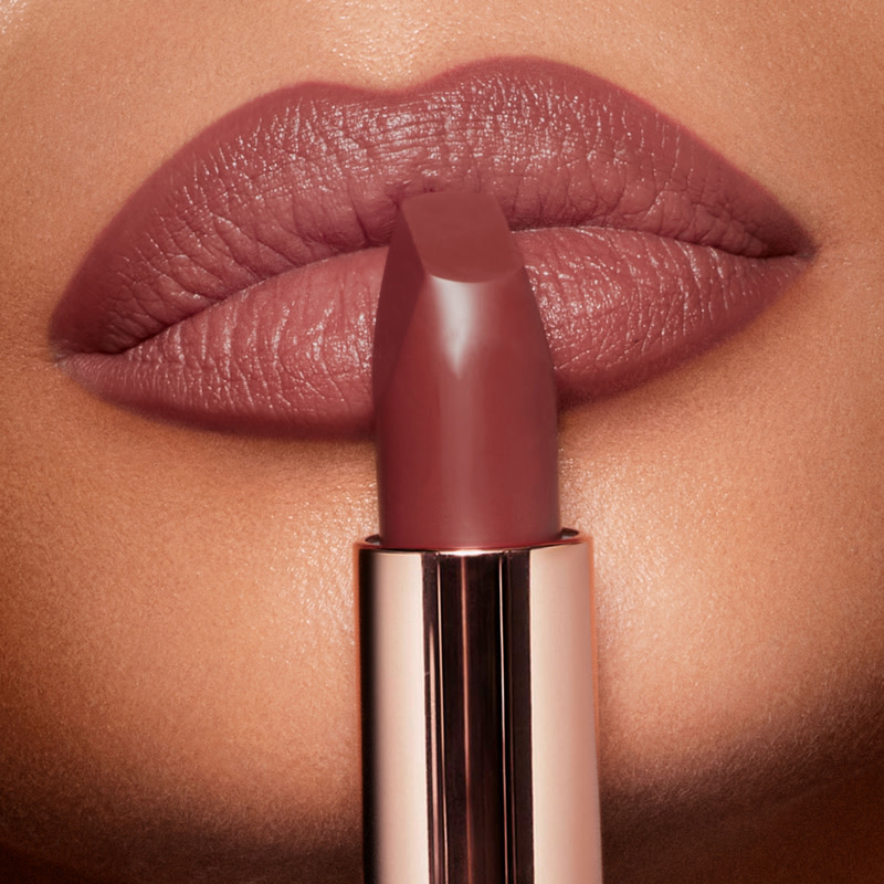 Labbra in primo piano di una modella dalla carnagione molto scura che indossa un rossetto opaco rosa berry e tiene il rossetto aperto davanti alle labbra. 