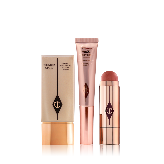 A light-beige-coloured primer in a rectangular bottle with a golden-coloured lid, a highlighting wand in rose-gold packaging, and an open blush stick in a terracotta colour in a reflective, golden-coloured tube. 