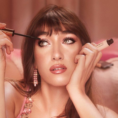 Light-tone brunette model with green eyes wearing nude pink lipstick with a satin finish while applying berry-brown, lengthening mascara that gives her lashes the appearance of false lashes.
