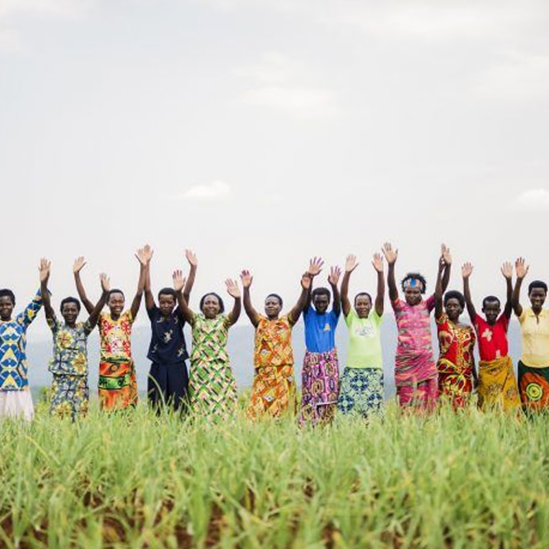 Deep-toned women wearing brightly coloured, geometric-patterned dresses while waving and smiling in a field. 