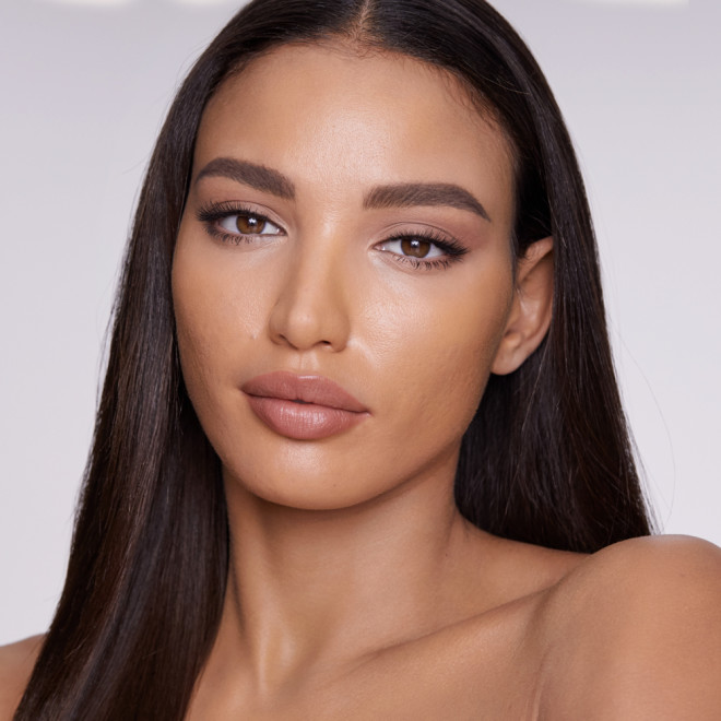 Medium-dark-tone model with flawless, matte face base, wearing nude pink matte lipstick with fawn eyeshadow, and black eyeliner.