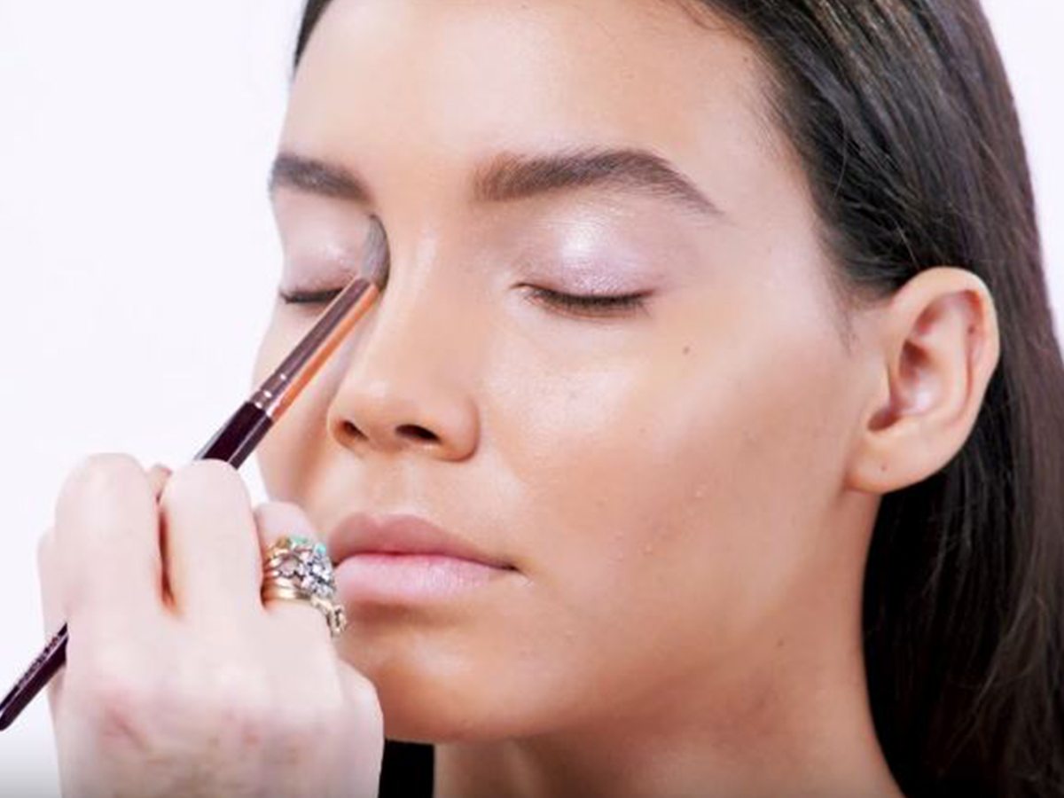 A light-tone brunette model with dewy skin getting a frosted lilac eyeshadow applied on her inner eye corner. 