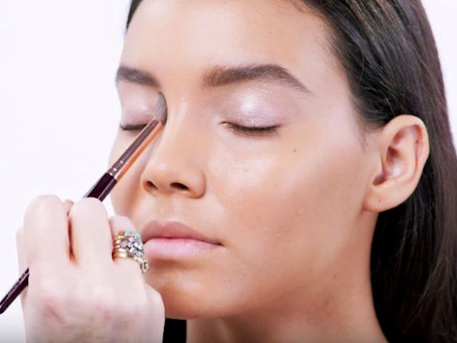 A light-tone brunette model with dewy skin getting a frosted lilac eyeshadow applied on her inner eye corner. 