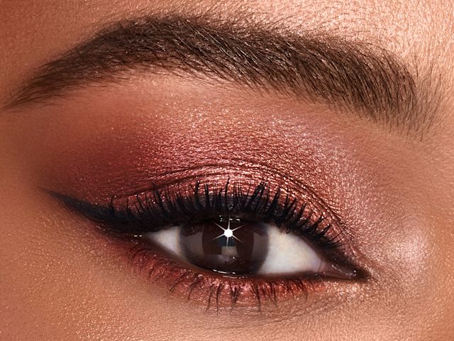 Single-eye close-up of a deep-tone model with brown eyes wearing shimmery rose gold and smokey brown eye shadow. 