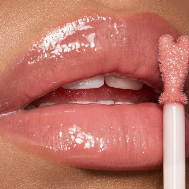 Lips close-up of a light-tone model applying sheer, nude pink lip gloss with a heart-shaped applicator. 