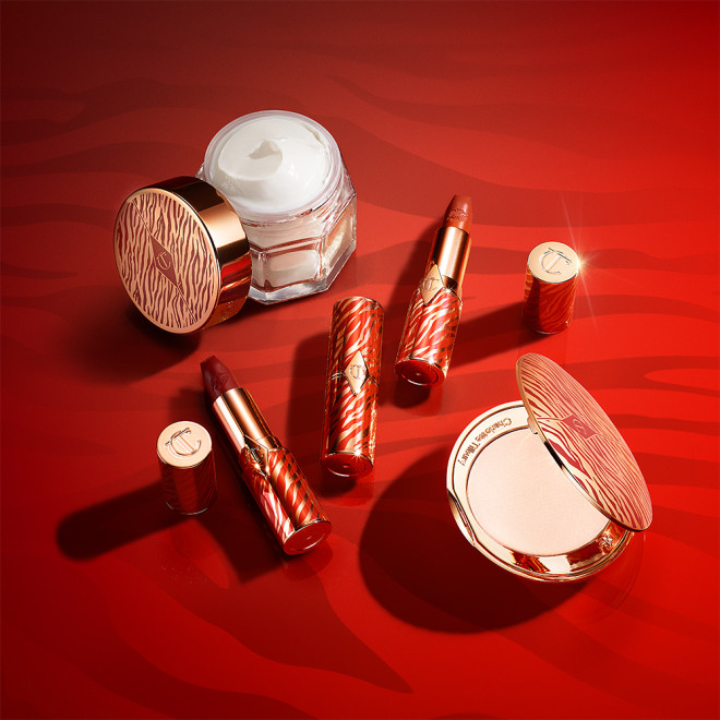 An open, face cream in a glass jar with a gold-coloured lid with three open lipsticks in shades of red in gold-coloured tubes, and open pressed powder compact in gold-coloured packaging with red stiger stripes on all of the products in celebration of the Lunar New Year.
