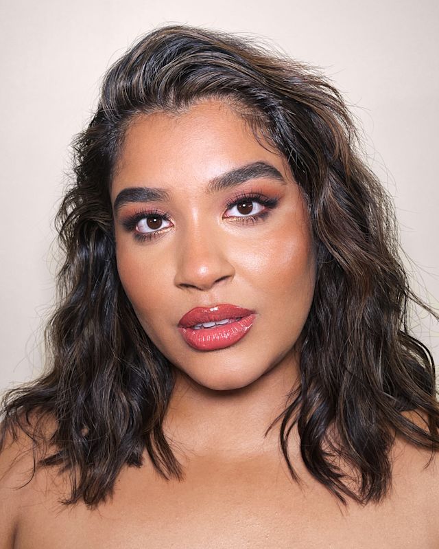Close-up of a medium-tone model with brown eyes wearing dark coral glossy lipstick with smokey brown eye makeup and glow peach blush.