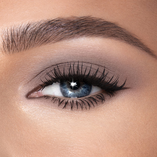 Single-eye close-up of a fair-tone model with blue eyes wearing smokey taupe cream eyeshadow smudged and spread on her eyelid with black eyeliner for a smokey taupe eye look. 