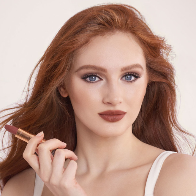 A fair-tone model wearing smokey eyeshadow with black eyeliner and a mid-toned muted nude-rose matte lipstick while holding the lipstick.
