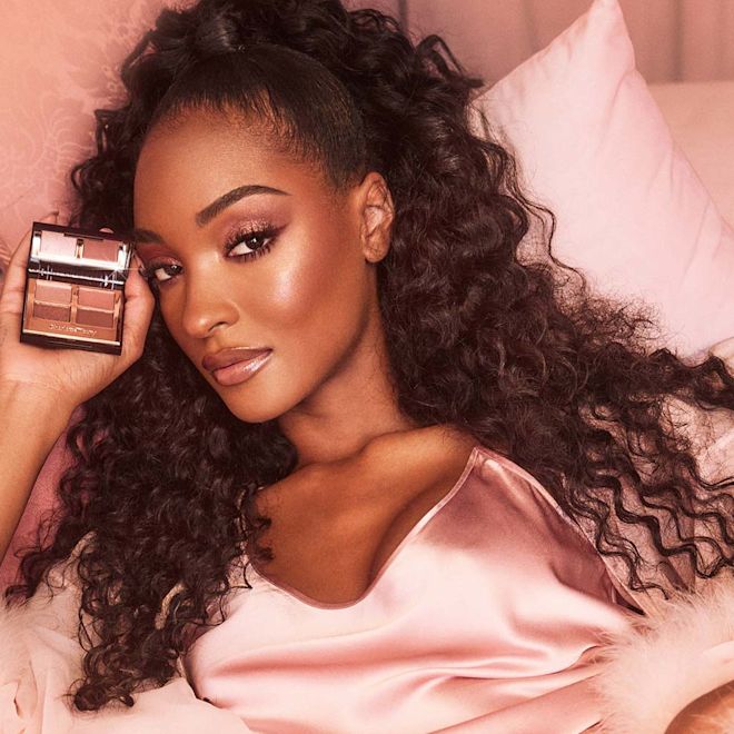 Pillow Talk Lux Palette Cat Box ES Deep-tone model with brown eyes wearing berry pink lipstick with a gloss on top with eye makeup in shades of pearlescent rose gold, dusky rose, berry brown and rose-bud pink while holding up a quad eyeshadow palette used to create the eye look.