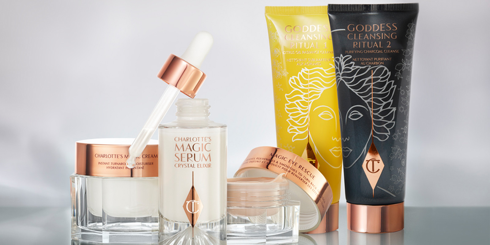 A pearly-white face cream in a glass jar and a dark champagne-coloured eye cream in a glass pot with a luminous face serum with its dropper lid removed, and two facial cleansers, one in lemon-yellow and the other in charcoal-black packaging, all four with rose-gold-coloured lids. 