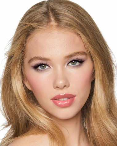 A light-tone model with green eyes wearing shimmery lilac and champagne eye makeup with nude pink blush and coral lipstick with gloss on top.