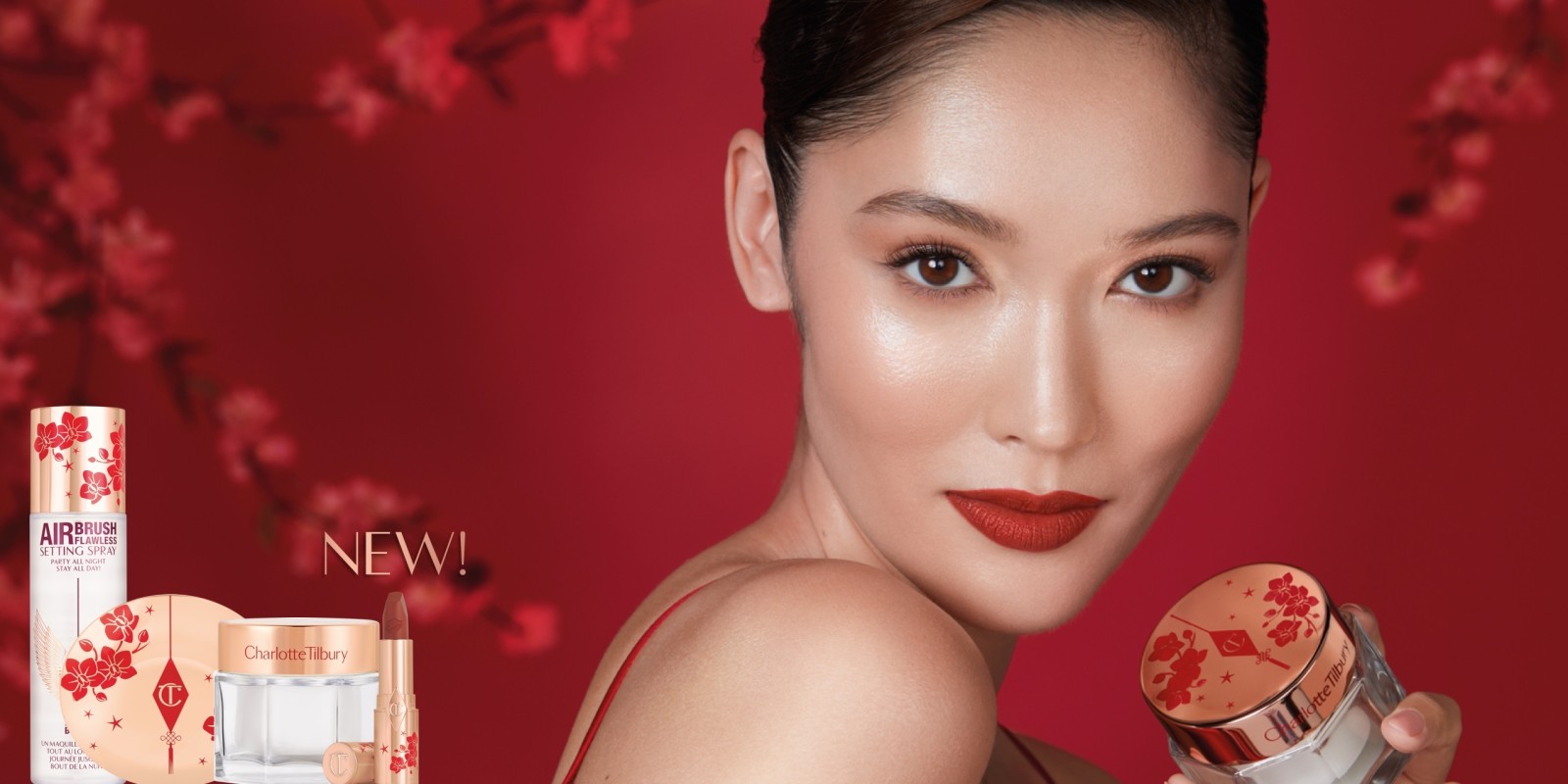 A fair skin model wearing matte burnt-orange red lipstick with rose gold shimmery eye makeup and holding a face cream with a gold-coloured lid with red cherry blossoms on it.