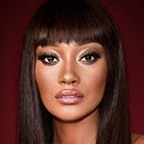 A deep-tone model with brown eyes wearing smokey green eye makeup with black eyeliner, soft brown blush, and nude pink lipstick topped up with gloss. 