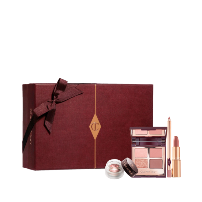 An open, mirrored-lid quad eyeshadow palette in matte and shimmery shades of pink and gold, an open matte lipstick in a nude pink shade, and crema eyeshadow in a glass pot with a dark brown lid, along with a white and scarlet makeup sleeve. 