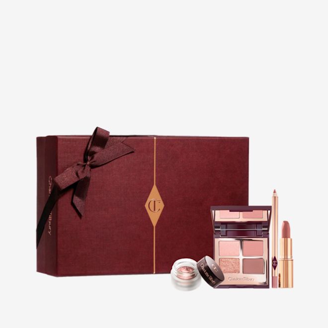 Pillow Talk Romance Box with Sleeve displayed An open, mirrored-lid quad eyeshadow palette in matte and shimmery shades of pink and gold, an open matte lipstick in a nude pink shade, and crema eyeshadow in a glass pot with a dark brown lid, along with a white and scarlet makeup sleeve.