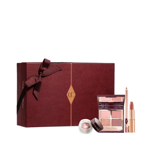 An open, mirrored-lid quad eyeshadow palette in matte and shimmery shades of pink and gold, an open matte lipstick in a nude pink shade, and crema eyeshadow in a glass pot with a dark brown lid, along with a white and scarlet makeup sleeve. 