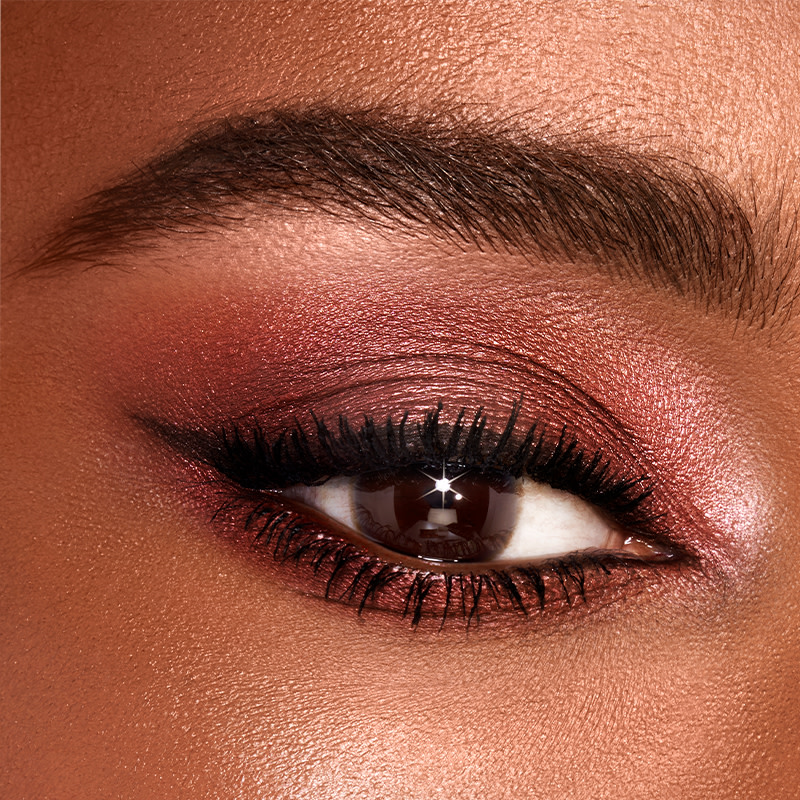 Single-eye close-up of a deep-tone model with brown eyes wearing shimmery red and rose-gold eyeshadow. 