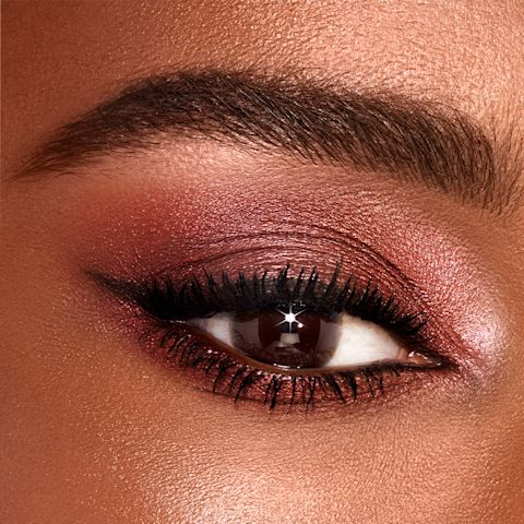Single-eye close-up of a deep-tone model with brown eyes wearing shimmery red and rose-gold eyeshadow. 
