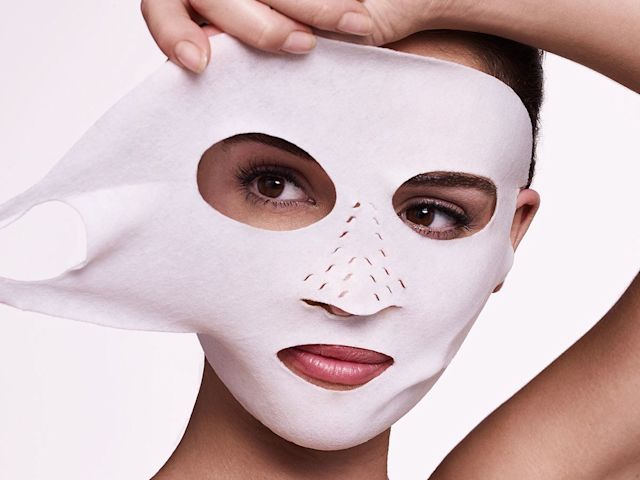 A medium-tone model applying a white sheet mask made of textile fabric with ear holes. 