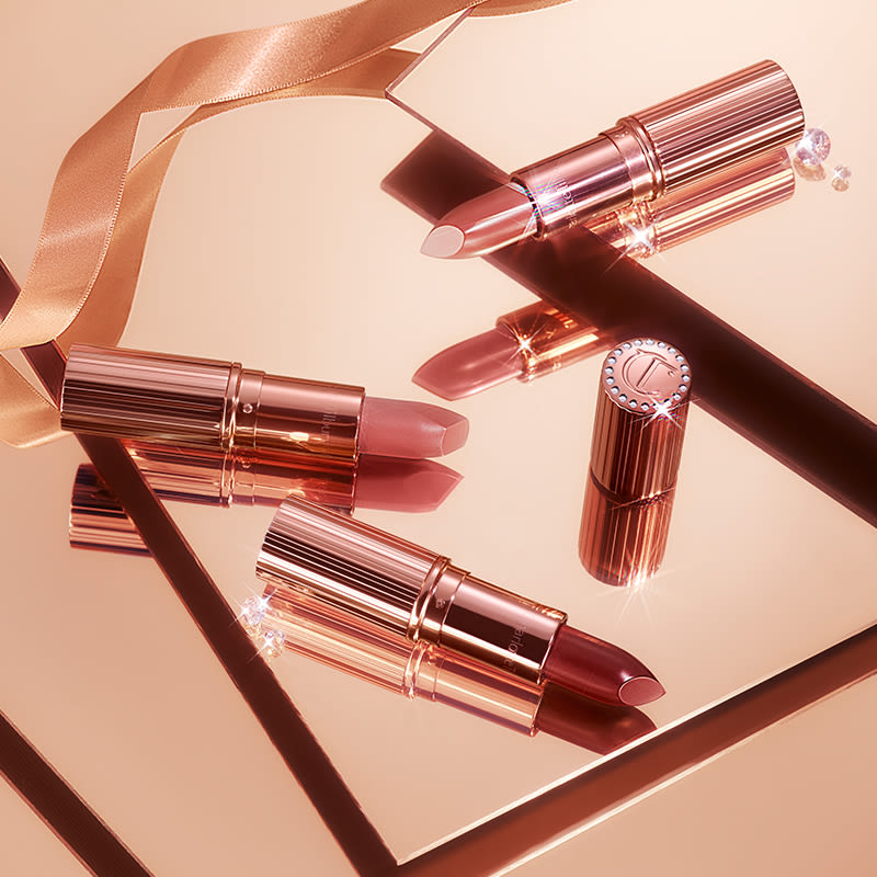 Three, open nude lipsticks in shades of nude peach, nude pink, and nude red placed on shiny, champagne-coloured tiles. 