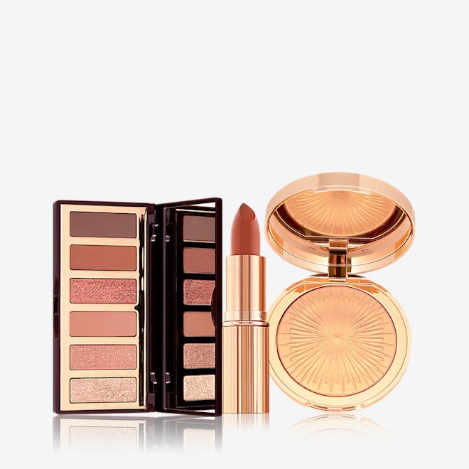Charlotte's Mesmerising Glow Kit open An open, 6-pan eyeshadow palette with a mirrored lid and matte and shimmery eyeshadows in shades of pink, pink, and brown, an open lipstick in a nude brick rose shade, and a highlighter compact in a soft gold shade.