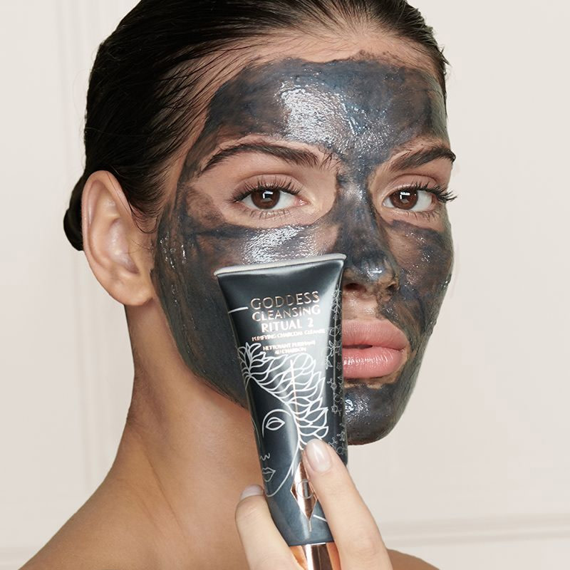 A light-tone model wearing a charcoal, wash-off mask cleanser while holding the cleanser in front of her. 