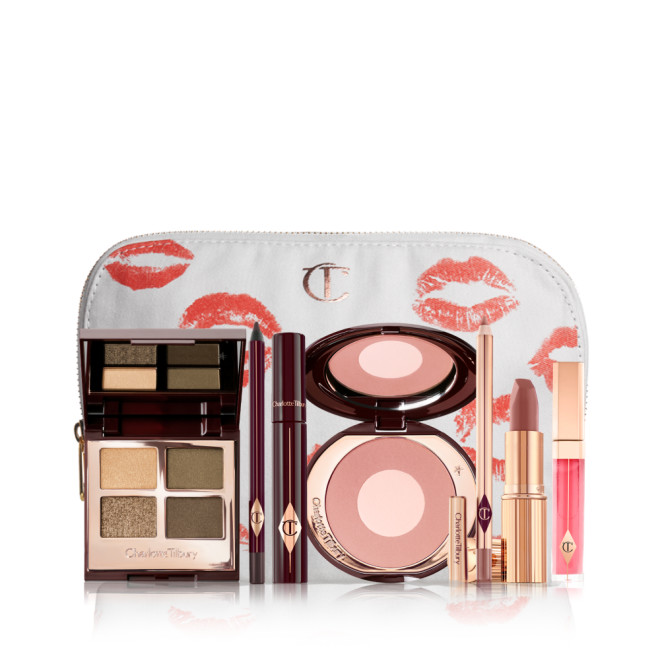 A white makeup bag with an open two-tone blush in cool-toned brown and warm pink with a mascara, eyeliner pencil, quad eyeshadow palette with shimmery and matte green and golden shades, an open lipstick in nude red-brown, lip liner pencil in taupe-brown, and a lip gloss in bright pink. 