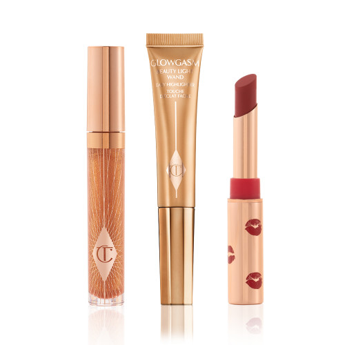 Lip gloss in a sheer gold colour in a glass tube with a gold-coloured lid, highlighter wand in a soft gold colour, and matte lipstick in a dark red shade in a gold-coloured tube with red kiss kiss-print pattern all over it.