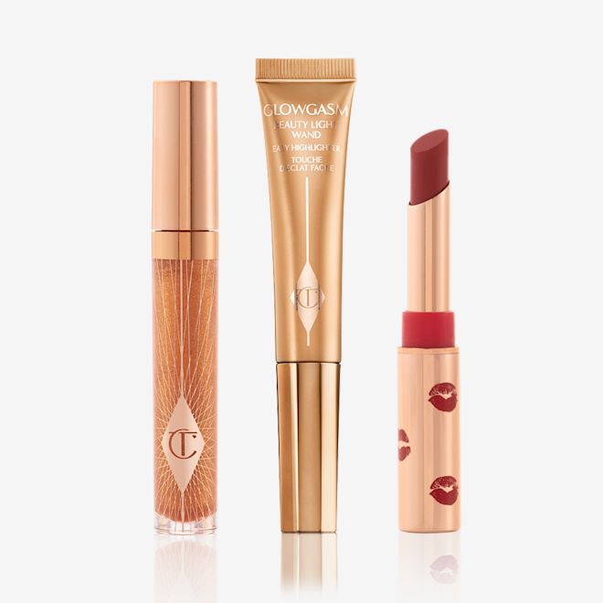 Glowing Golden Holiday Kit displayed Lip gloss in a sheer gold colour in a glass tube with a gold-coloured lid, highlighter wand in a soft gold colour, and matte lipstick in a dark red shade in a gold-coloured tube with red kiss kiss-print pattern all over it.