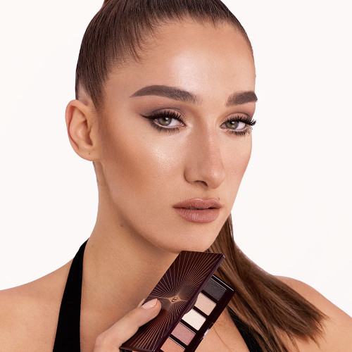 A fair-tone model with green eyes holding an open, mirrored-lid six-pan eyeshadow palette with matte eyeshadows in brown, peach, and beige shades while wearing a smokey brown eye look created using that palette with nude peach lipstick.