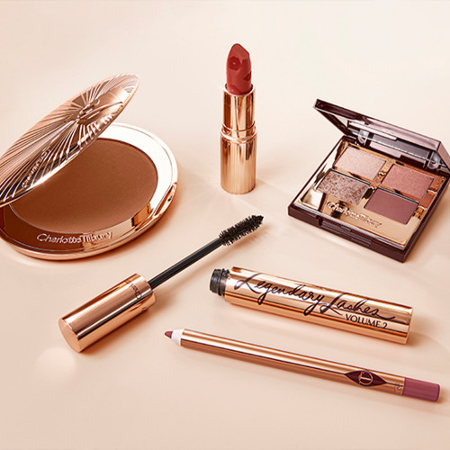 An open bronzer compact, open lipstick in a golden peachy-pink shade, quad eyeshadow palette with ivory-cream, rose-gold, red-brown and transparent sparkle shades, open mascara, and a deep nude pink lip liner pencil.  