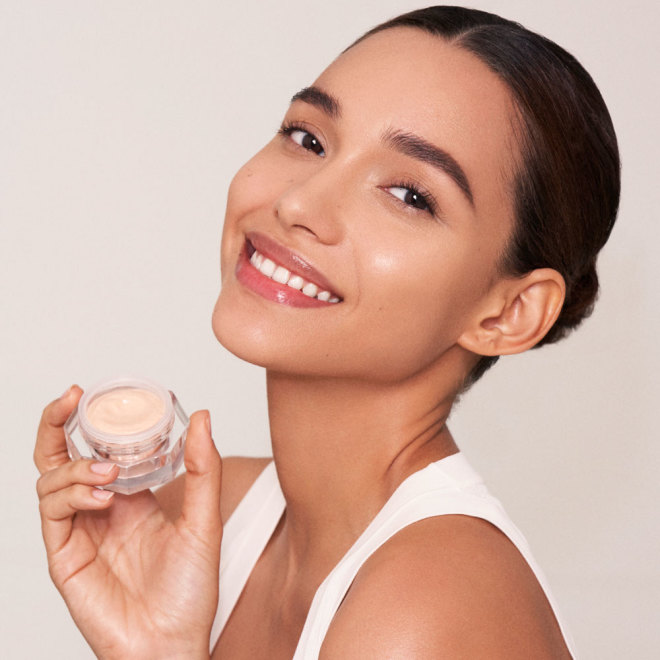 A light-tone brunette model with flawless, glowy, glass skin holding a petite jar of a light fawn-coloured eye cream.