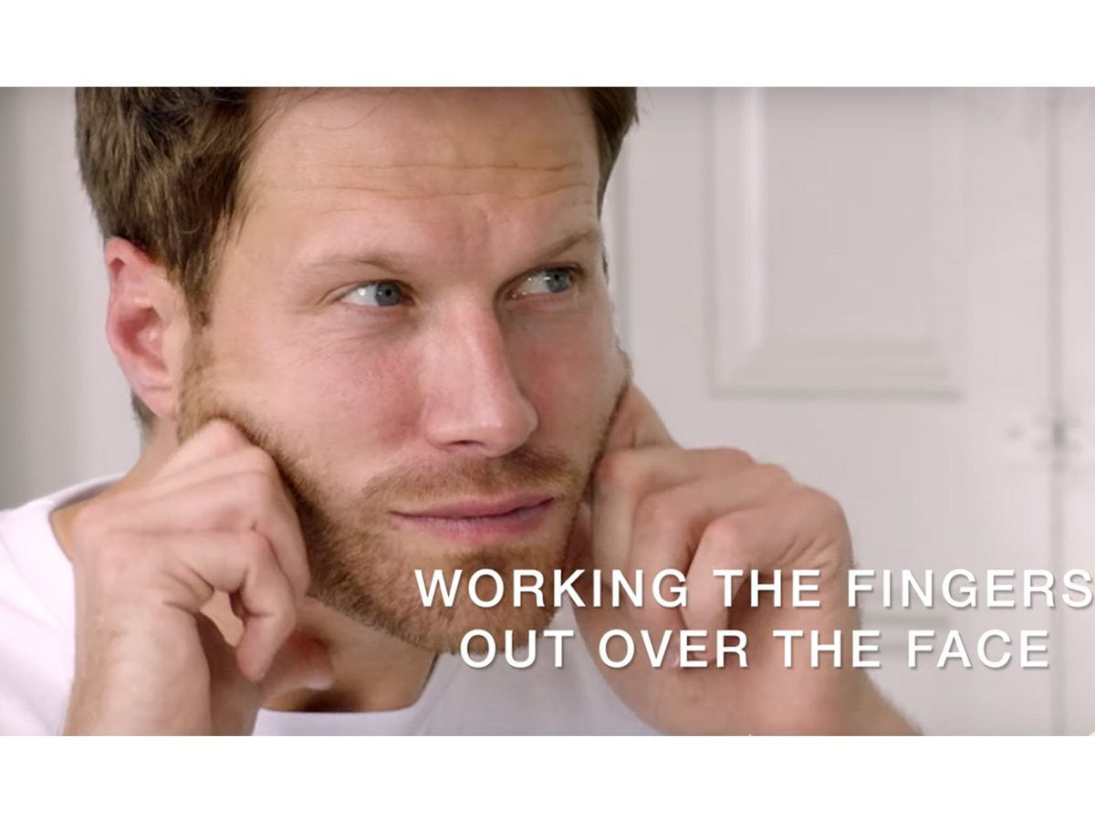 A light-tone, brunette male model massages a face cream into the skin with his fingertips.