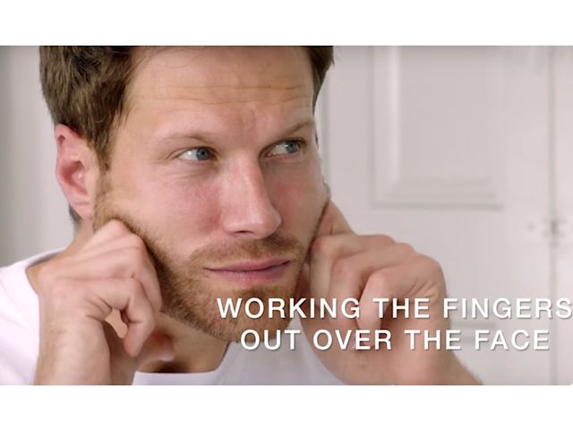 A light-tone, brunette male model massages a face cream into the skin with his fingertips.