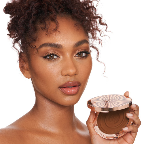 Deep-tone brunette model with brown eyes wearing nude pink lip gloss with glowy, cream bronzed for a sculpted yet natural makeup look while holding up an open bronzer compact.