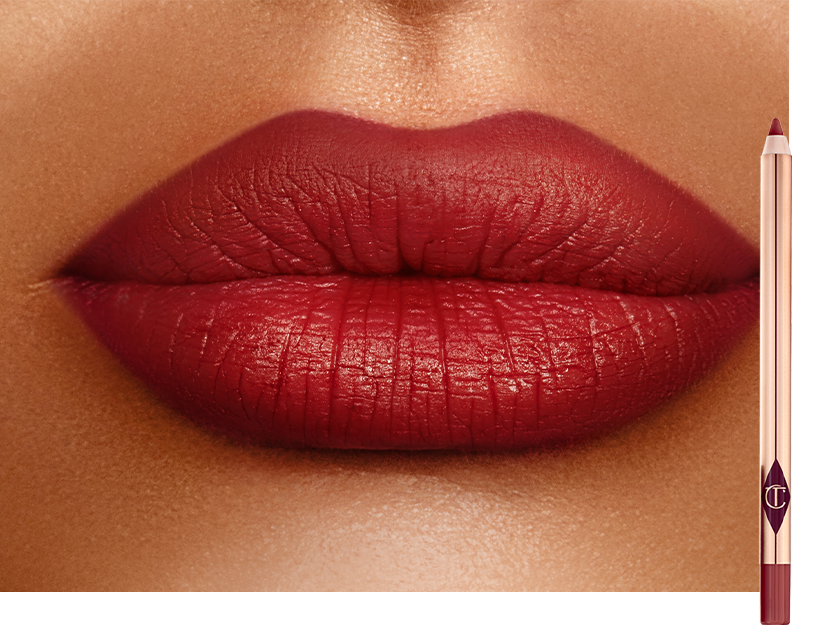 Lips close-up of a deep-tone model wearing a blood-red lip liner pencil all over her lips with the lip liner pencil next to her. 