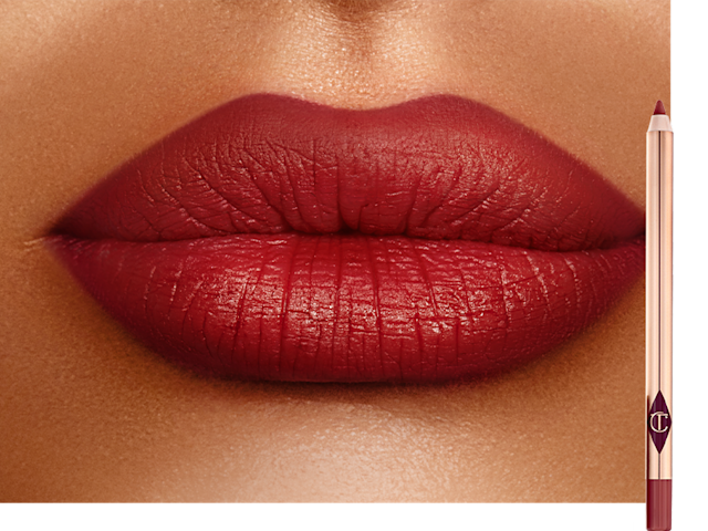 Lips close-up of a deep-tone model wearing a blood-red lip liner pencil all over her lips with the lip liner pencil next to her. 