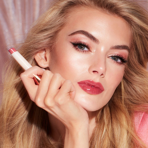 Fair-tone model with blue eyes wearing a moisturising lipstick balm in a peach rose shade with a high-shine finish while holding the lip balm.