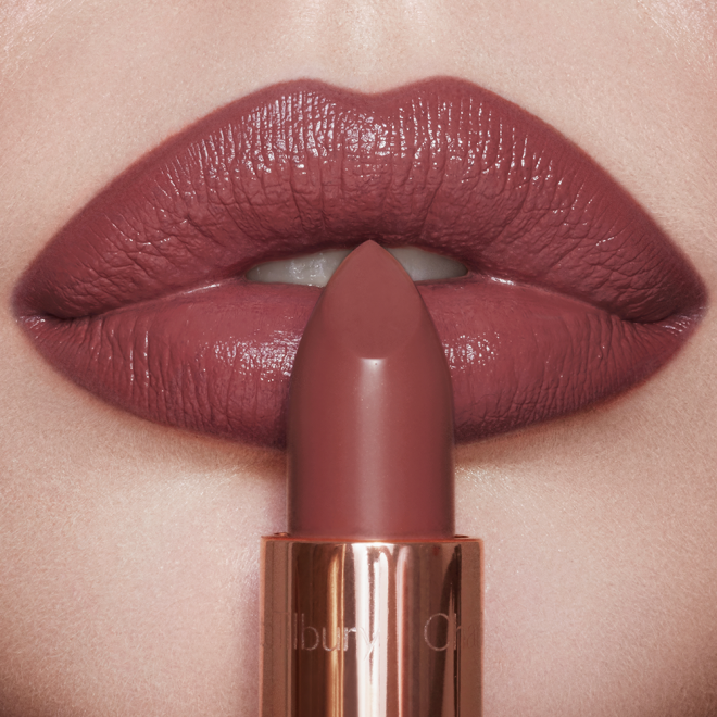 Lips close-up of a light-tone model wearing a dark, brown-pink lipstick with a satin finish while holding the lipstick in front of her.  