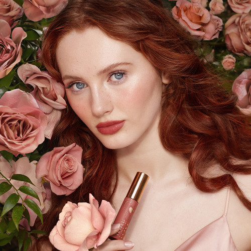 A fair-tone model with blue eyes wearing shimmery beige eyeshadow with a beachy nude-brown lip tint and holding the lip tint in one hand and a flower in the other.