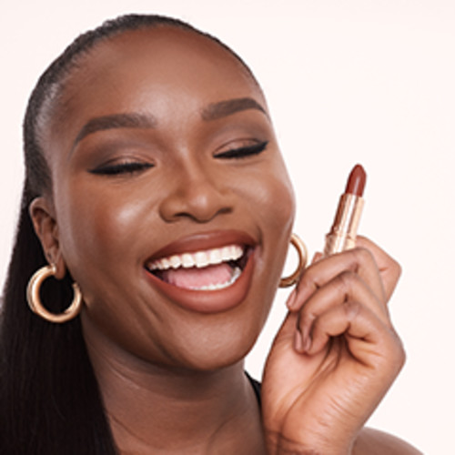 A deep-tone model wearing a deep, sultry rose-brown nude lipstick with a matte finish while holding that lipstick up. 