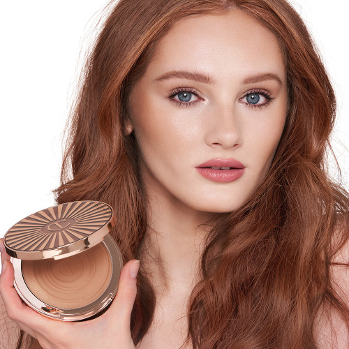 Fair-tone auburn-hair model with blue eyes wearing nude pink lip gloss with glowy, cream bronzed for a sculpted yet natural makeup look while holding up an open bronzer compact.