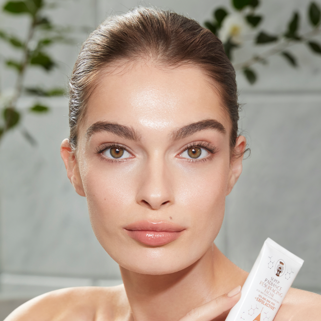 Light-tone model with hazel eyes and smooth, flawless, and glowy glass skin holding a white-coloured tube of a wash-off exfoliating mask.