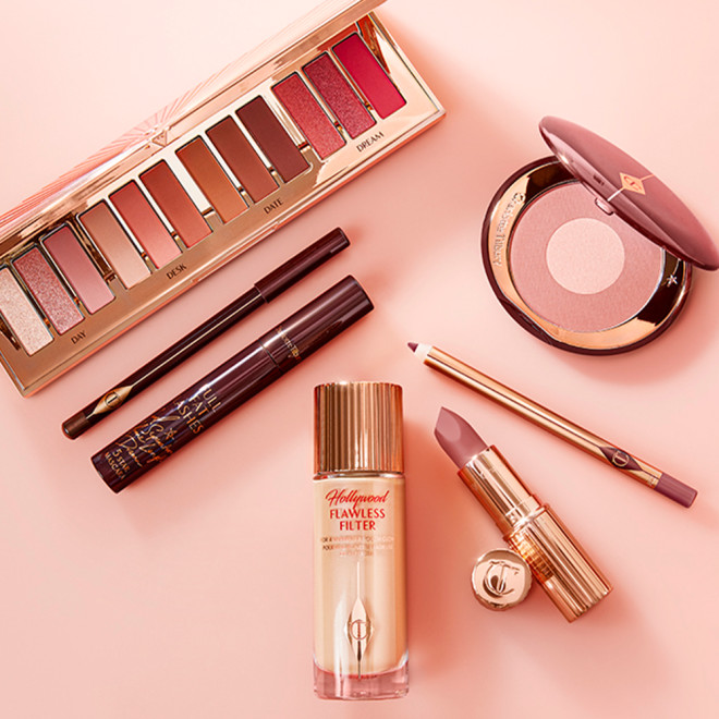 An open eyeshadow palette in pink, peach, gold, and brown shades, luminous primer in a glass bottle, black mascara and eyeliner, two-tone blush in light pink, and lip liner and lipstick in nude pink. 